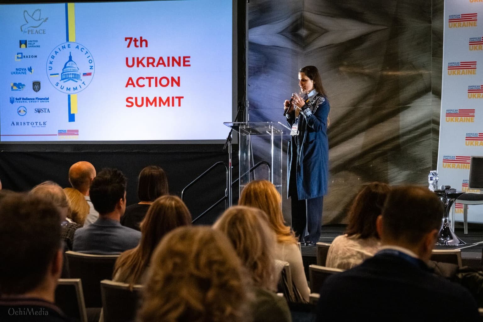 A speaker stands at a podium facing an audience, standing next to a Ukraine Action Summit presentation.