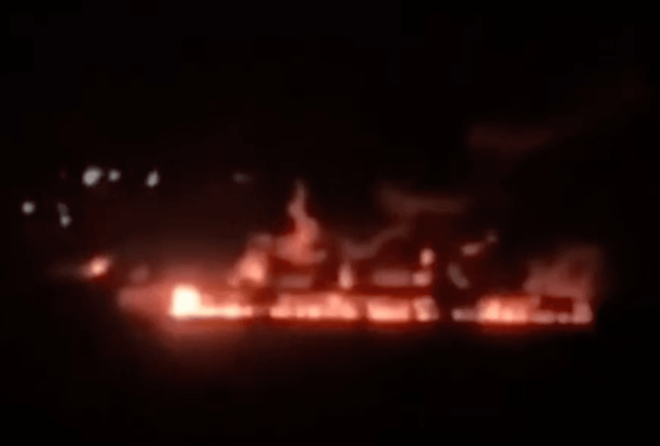 A fire burns at an industrial city under a black sky