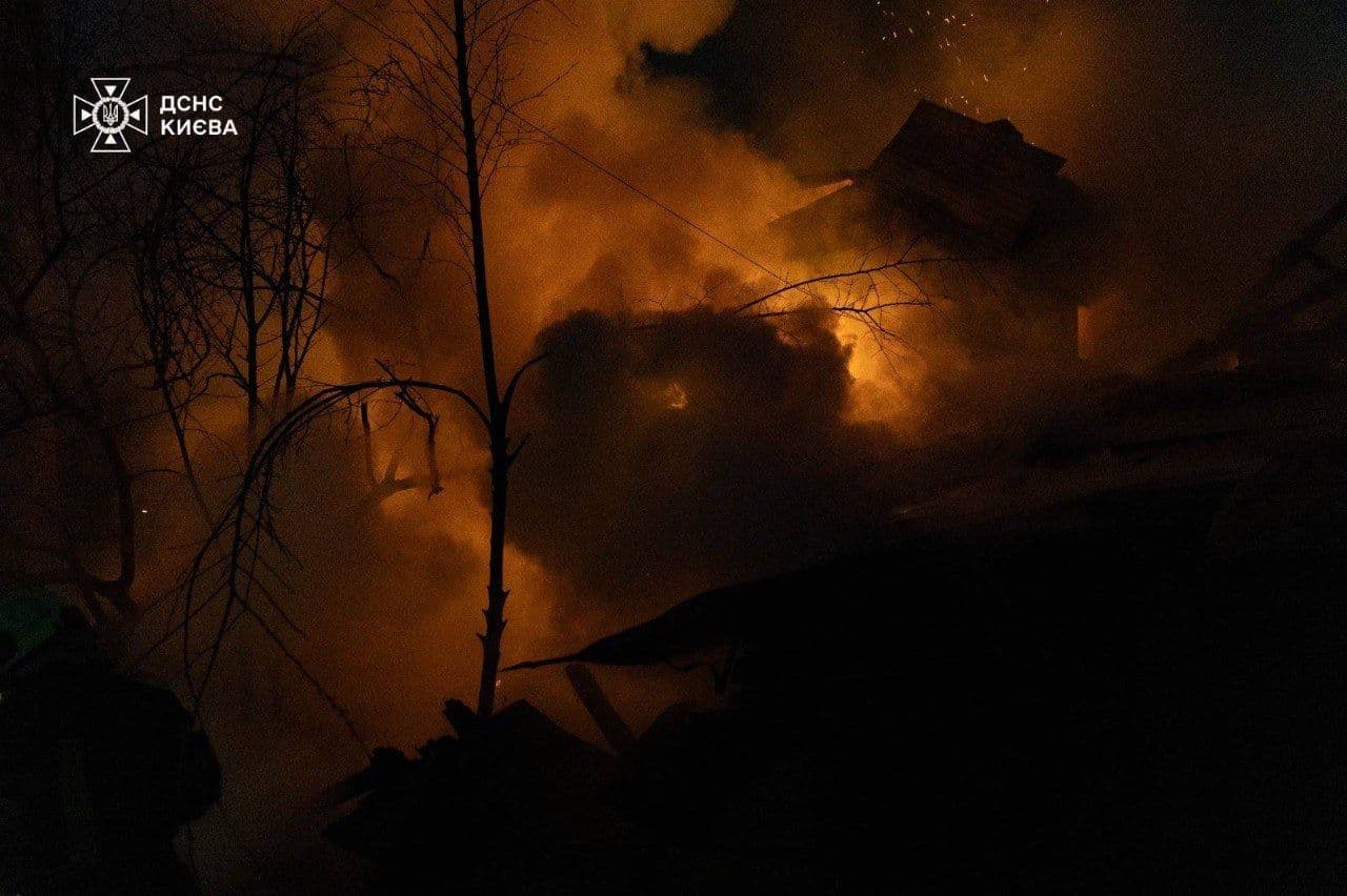 A bright orange fire burns behind dark smoke, the silhouette of a tree, and a house.