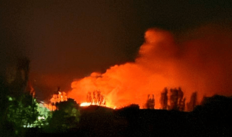 A large fire burns behind a treeline under a night sky