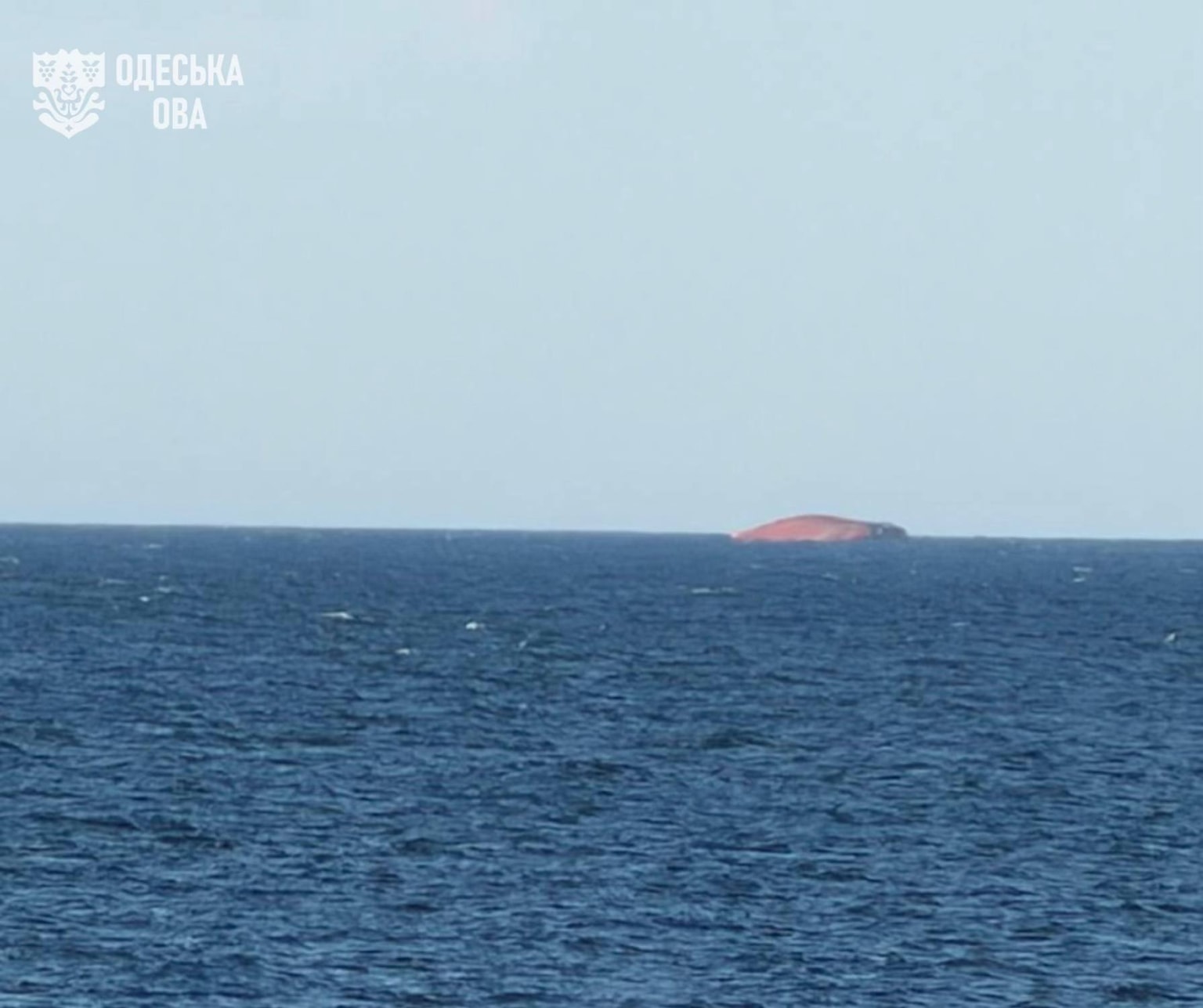 A capsized cargo vessel with an orange/red belly and dark hull elsewhere floats on a deep blue sea